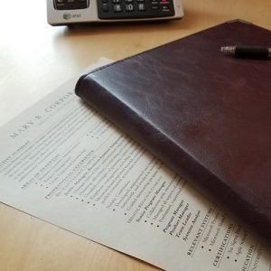 Image of a resume lying partially obscured by a portfolio with a black pen lying on top of it, on a light wood desk next to a business phone